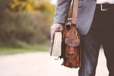 Un hombre sostiene un libro en su mano derecha junto a la cartera que cuelga de su hombro